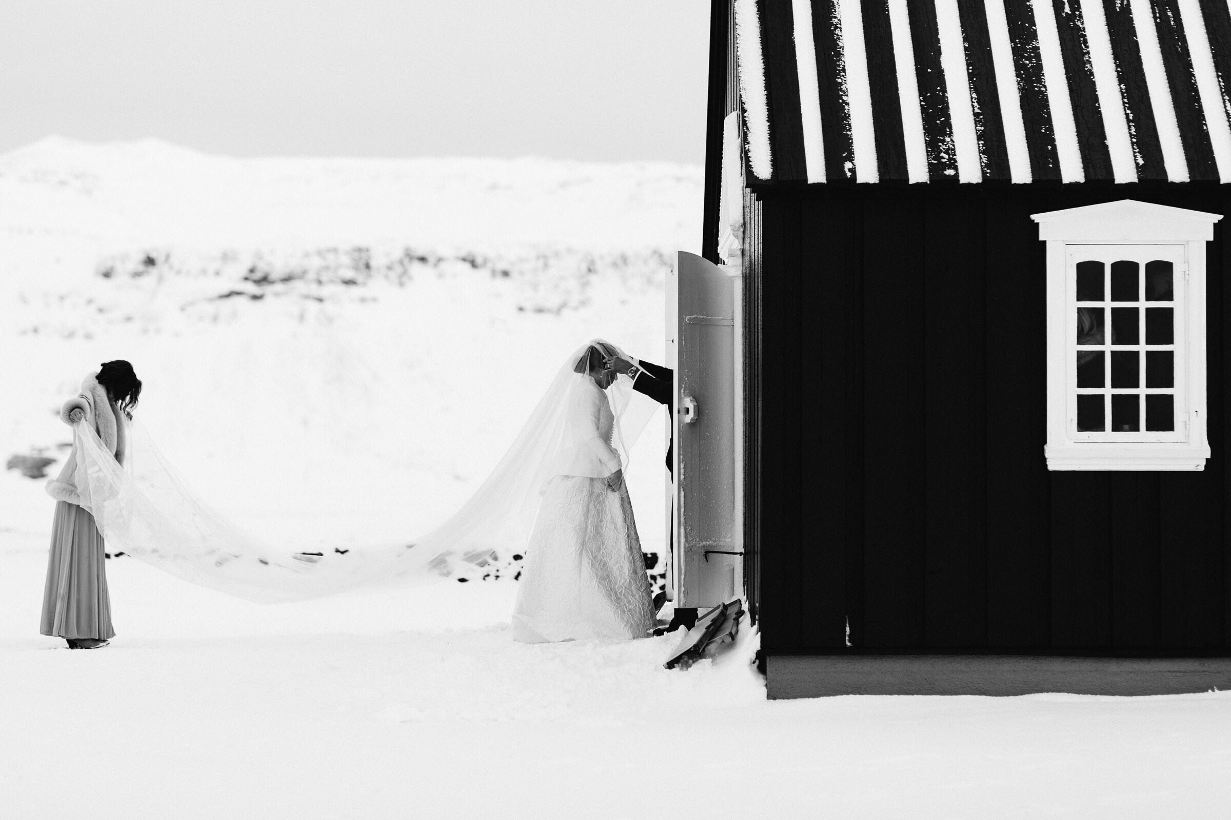 A bride standing out side a black church in the snow. Having her veil fixed before she enters her Iceland Destination wedding ceremony.