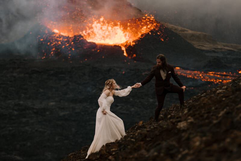 A wedding couple hike up a hill in their wedding outfits with an active volcano behind them.