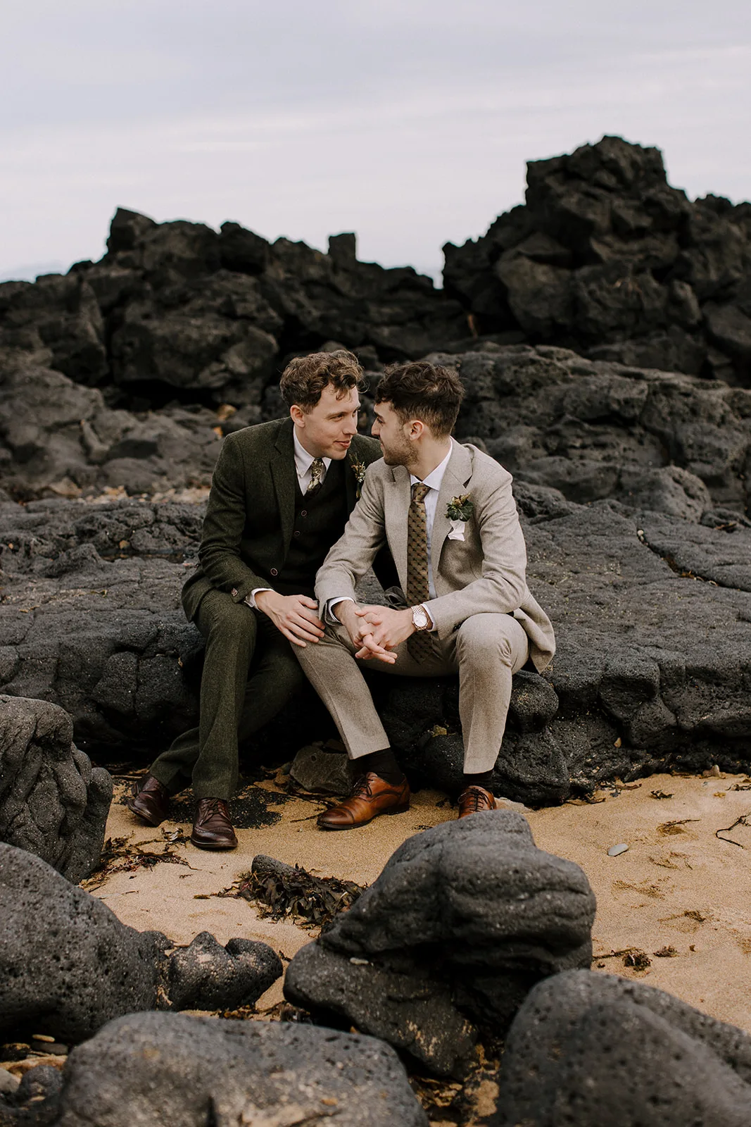 Two grooms sitting on the beach, looking each other in the eyes. Dressed in wool suits.
