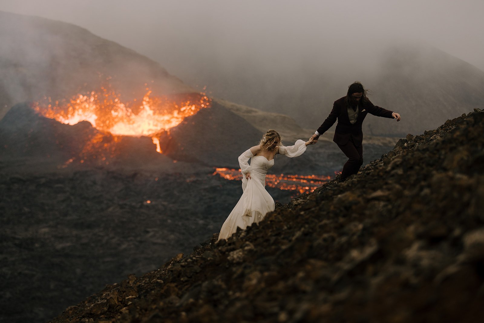 A bride and groom climb a hill in front of an active volcano on their Iceland elopement day.