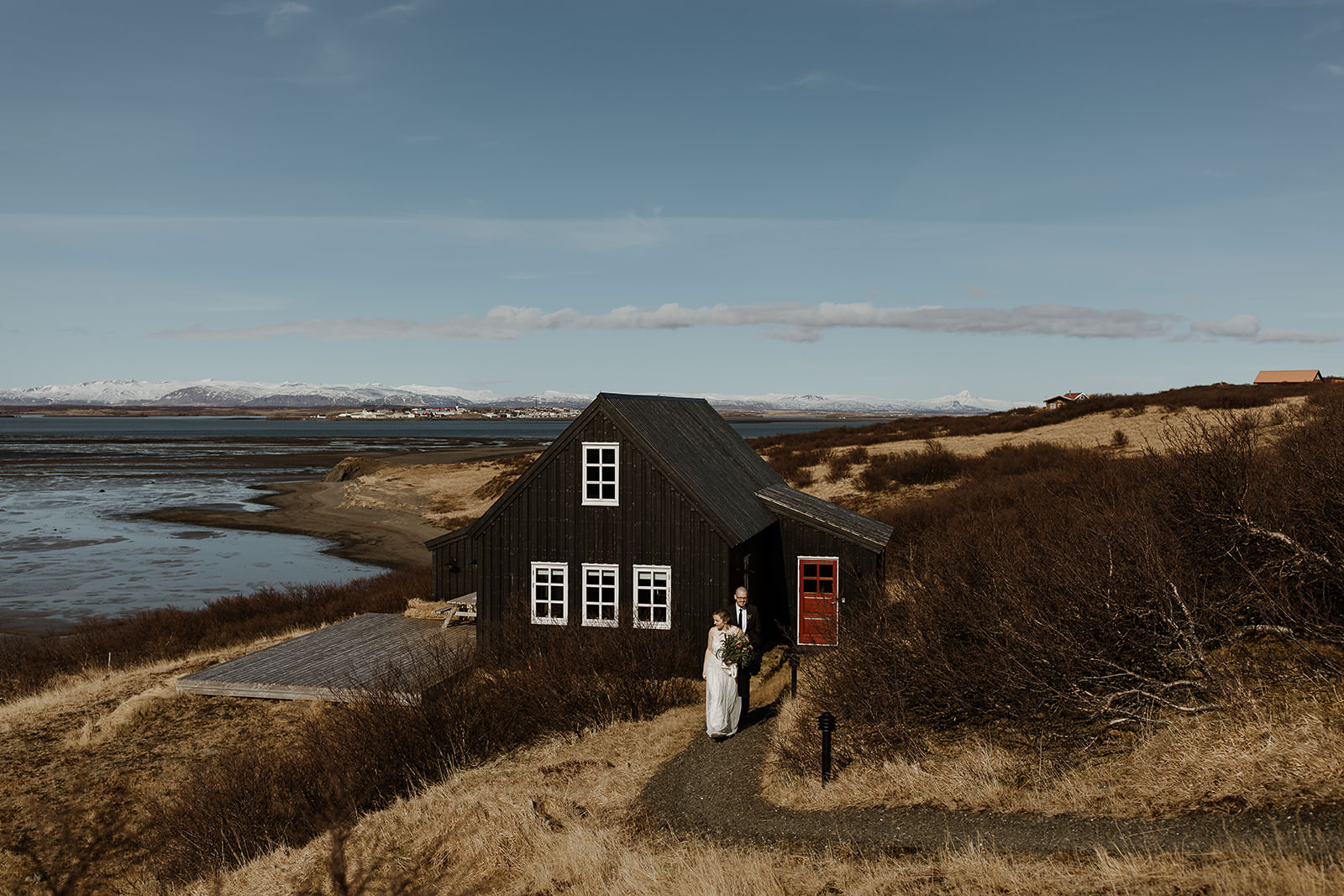 A wedding couple walks from an old black timber house on the west coast of Iceland.