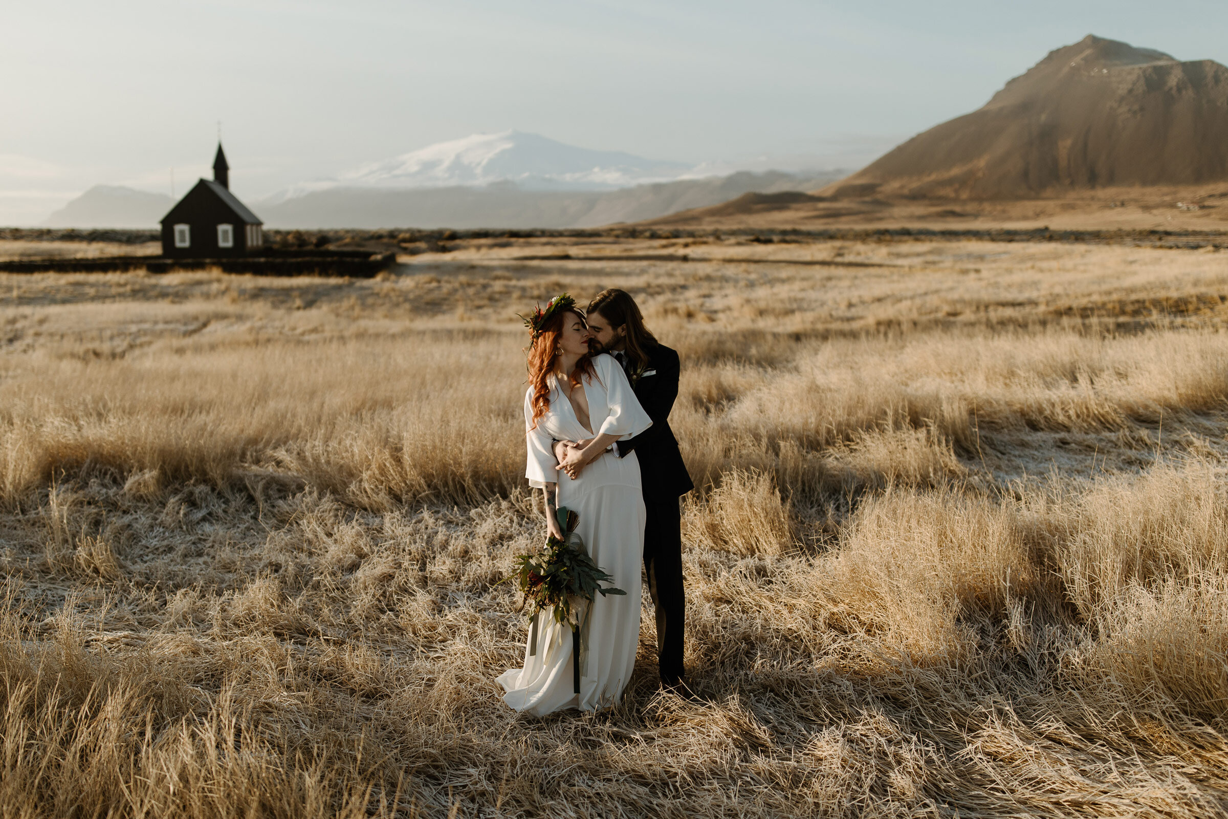 A stylish wedding couple embrace in at the back of the Búðir church with Snæfellsjökull glacier in the background.