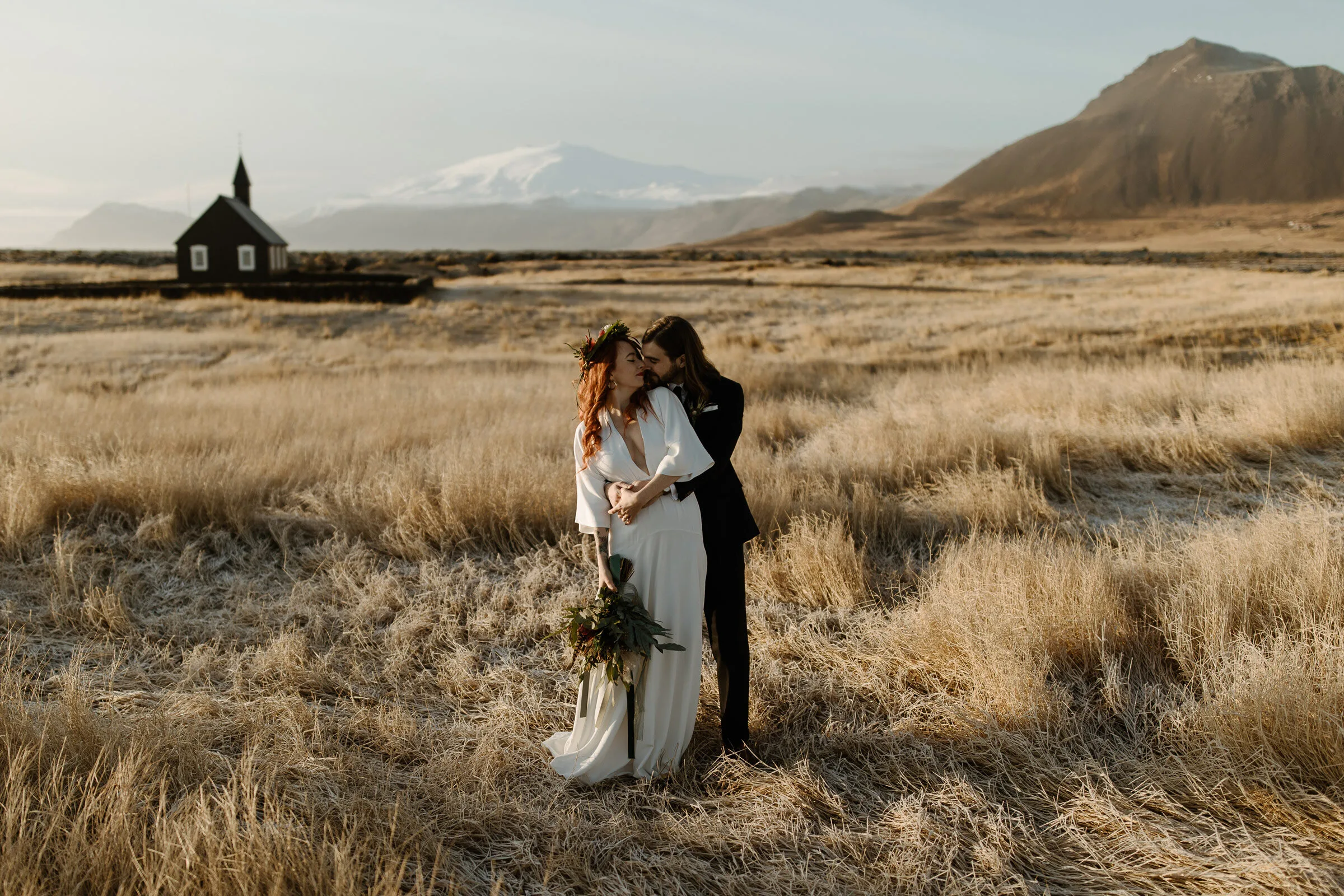 A stylish wedding couple embrace in at the back of the Búðir church with Snæfellsjökull glacier in the background.