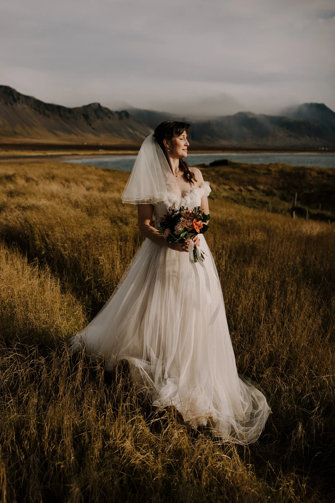 A bride standing in a field of yellow grass, eyes closed, facing the sun, taking in the moment.