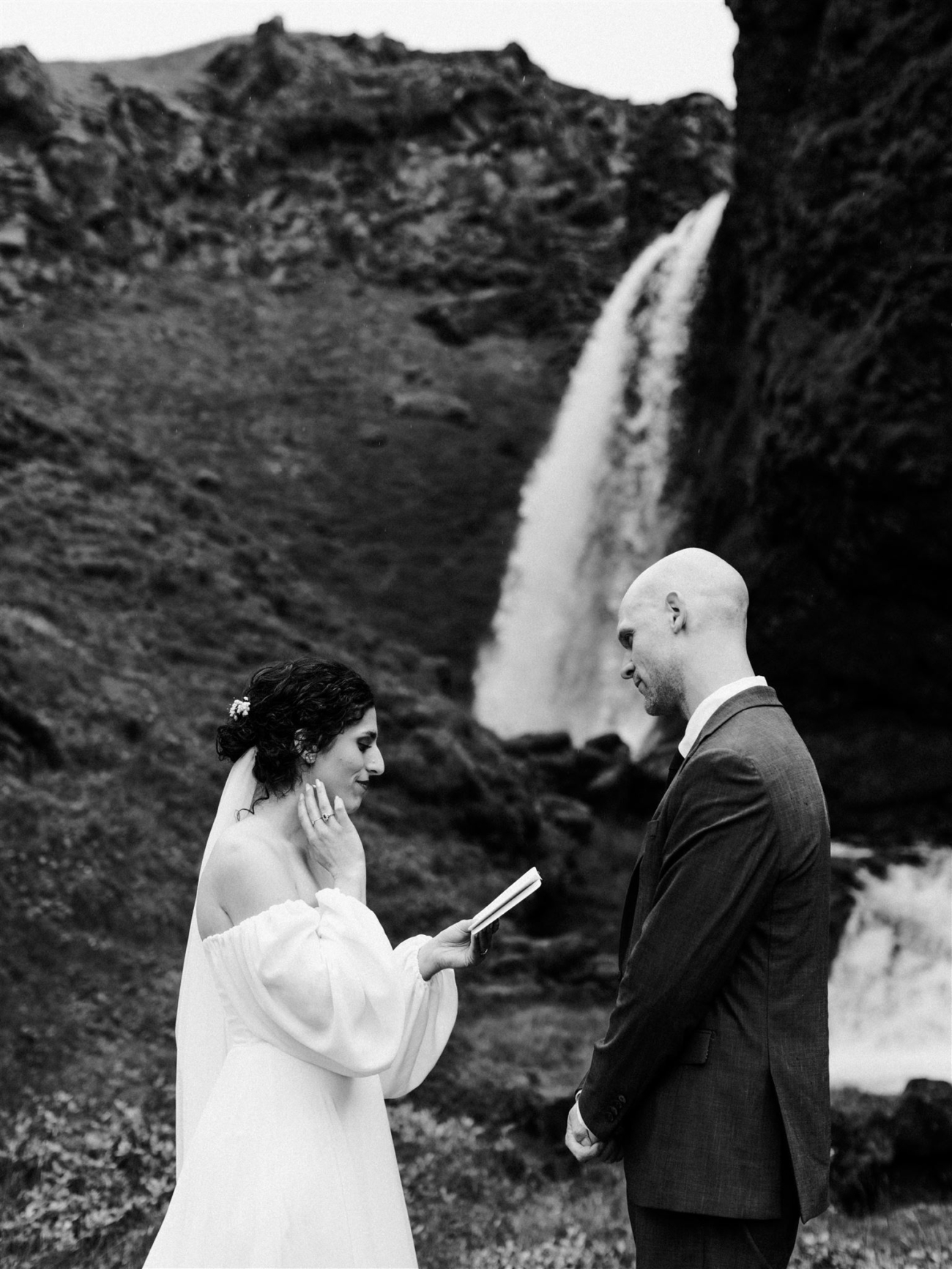 A bride reads her vows from a beautiful vow book as a waterfall thunders behind her in the distance during an Iceland elopement day.
