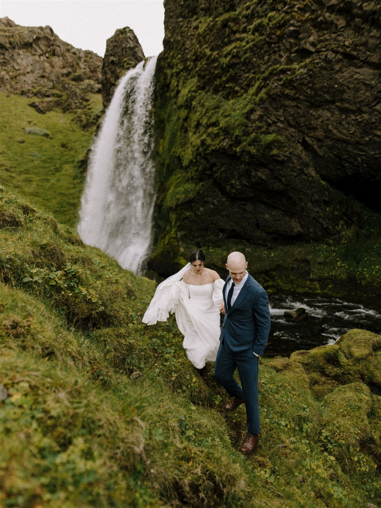 A wedding couple hiking in canyon with a roaring waterfall in the background on their elopement day in Iceland.