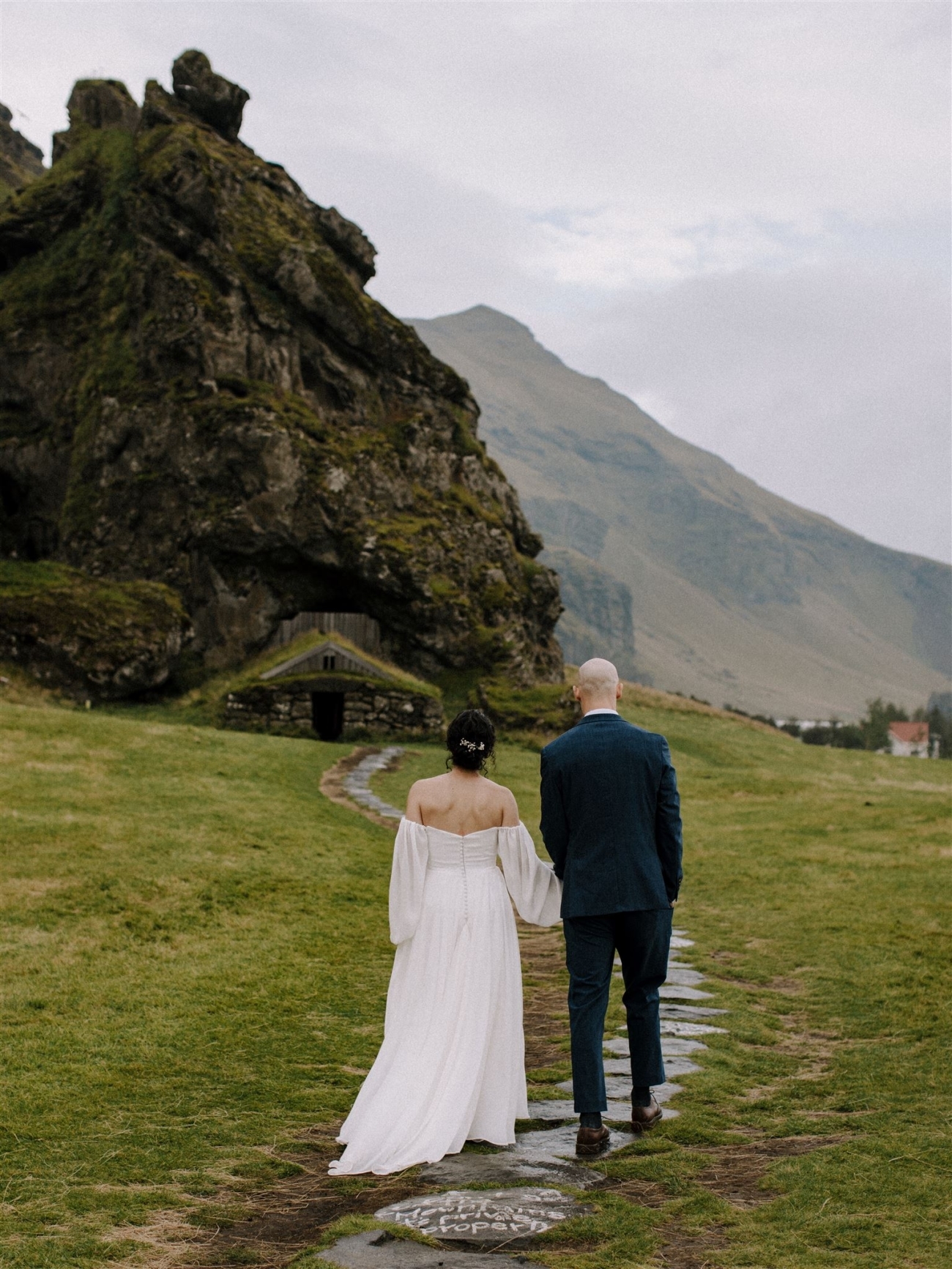 A bride and groom walk along a path towards an old turf house on a moody, overcast day on the south coast of Iceland.