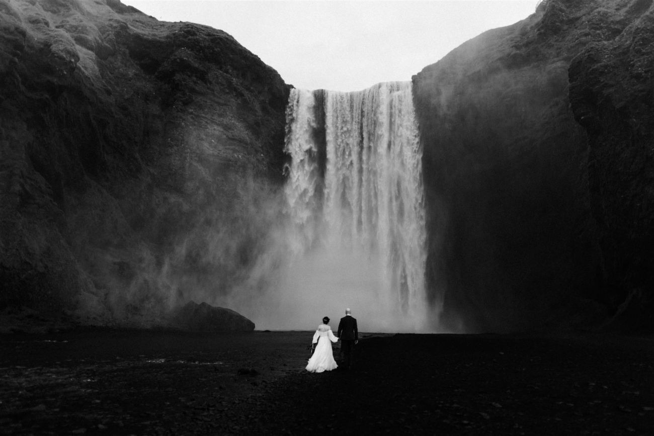 A wide shot of a wedding couple walking in the mist towards the thundering Skógafoss waterfall.