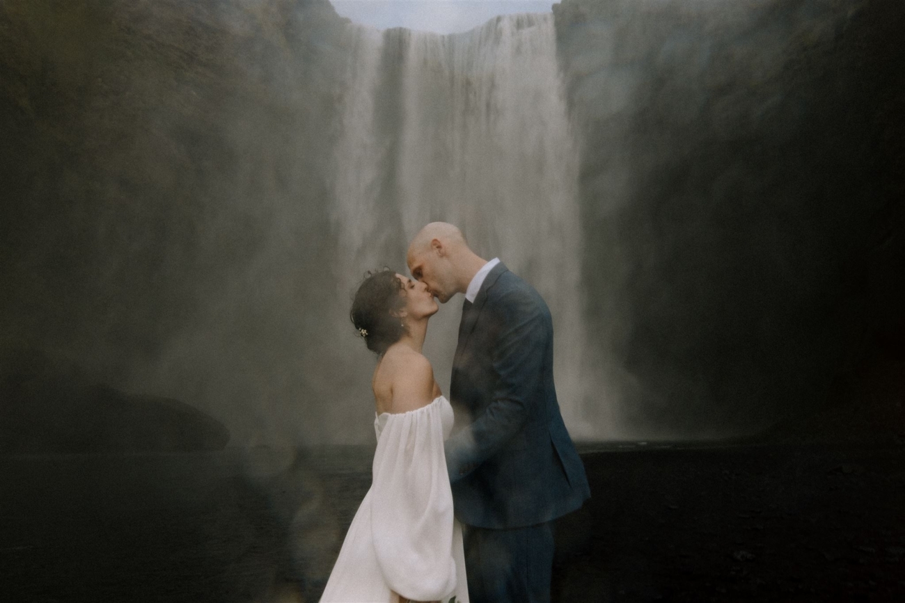 A bride and groom kissing and getting soaked in the mist of the thundering Skógafoss waterfall on their Iceland elopement day.