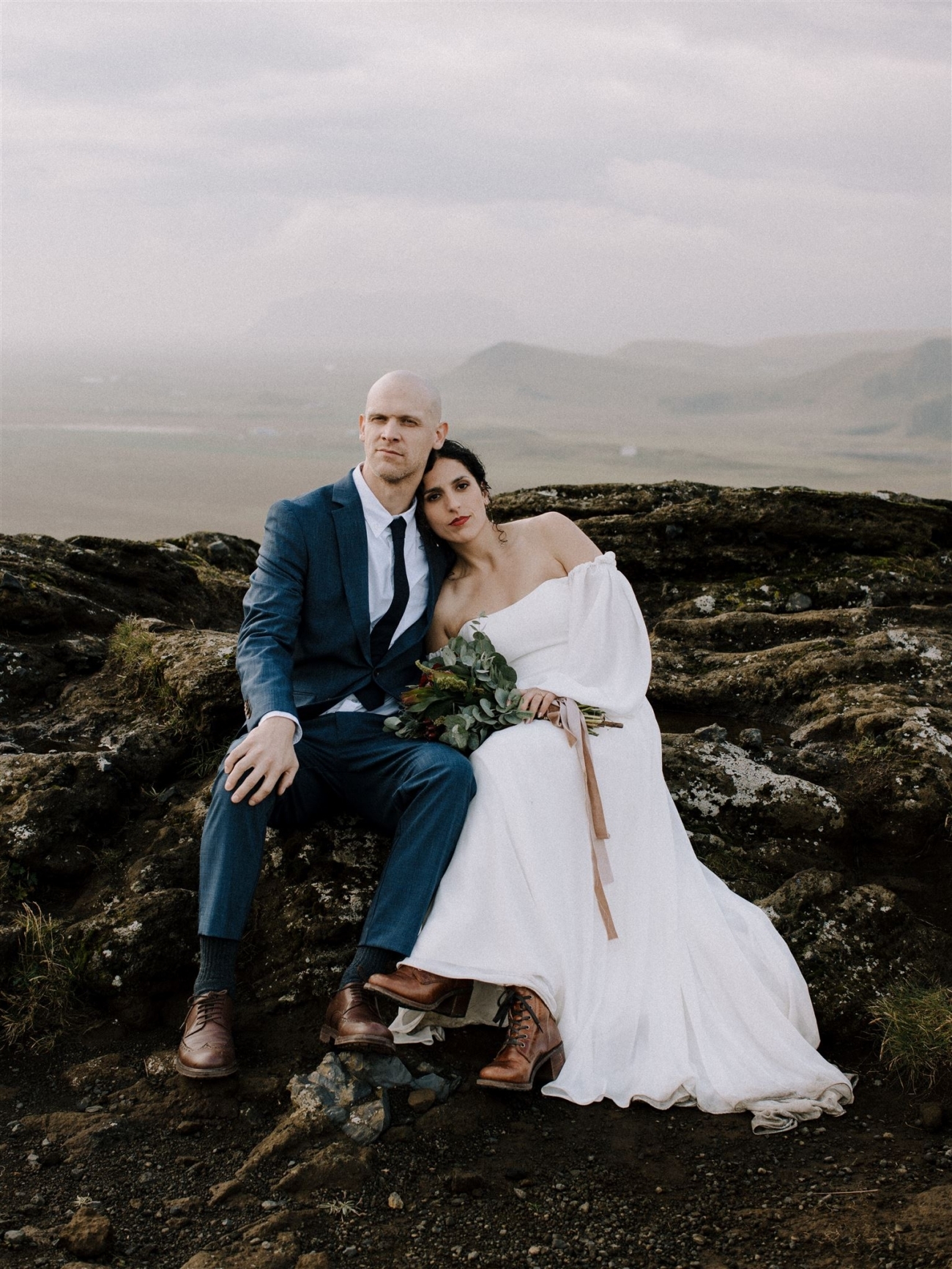 A classic portrait of a groom and bride sitting on some lava rocks on the south coast of Iceland. They both look calmly into the lens.