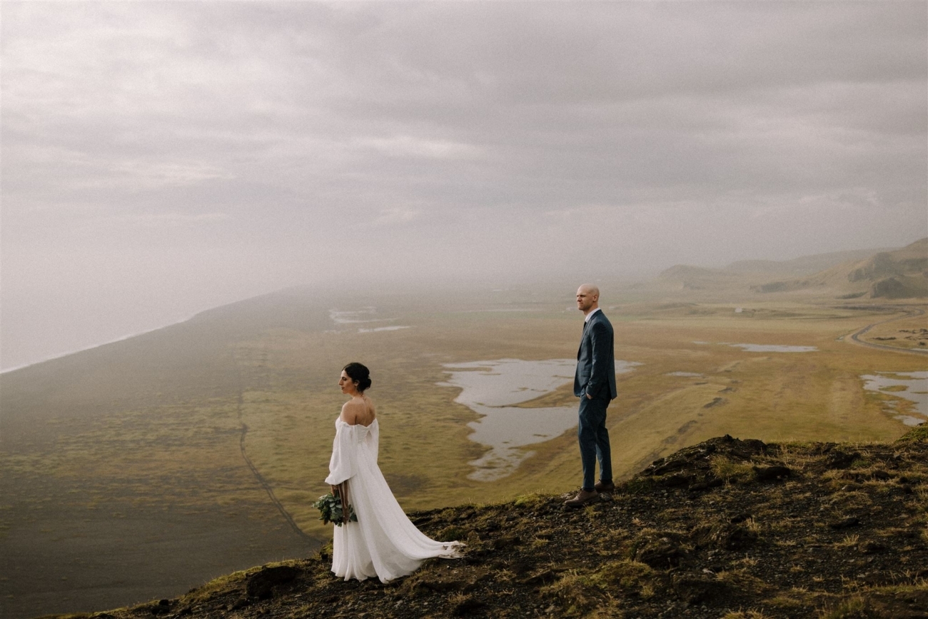 A bride and groom standing at the edge of Dyrhólaey cliffs staring into the distance.