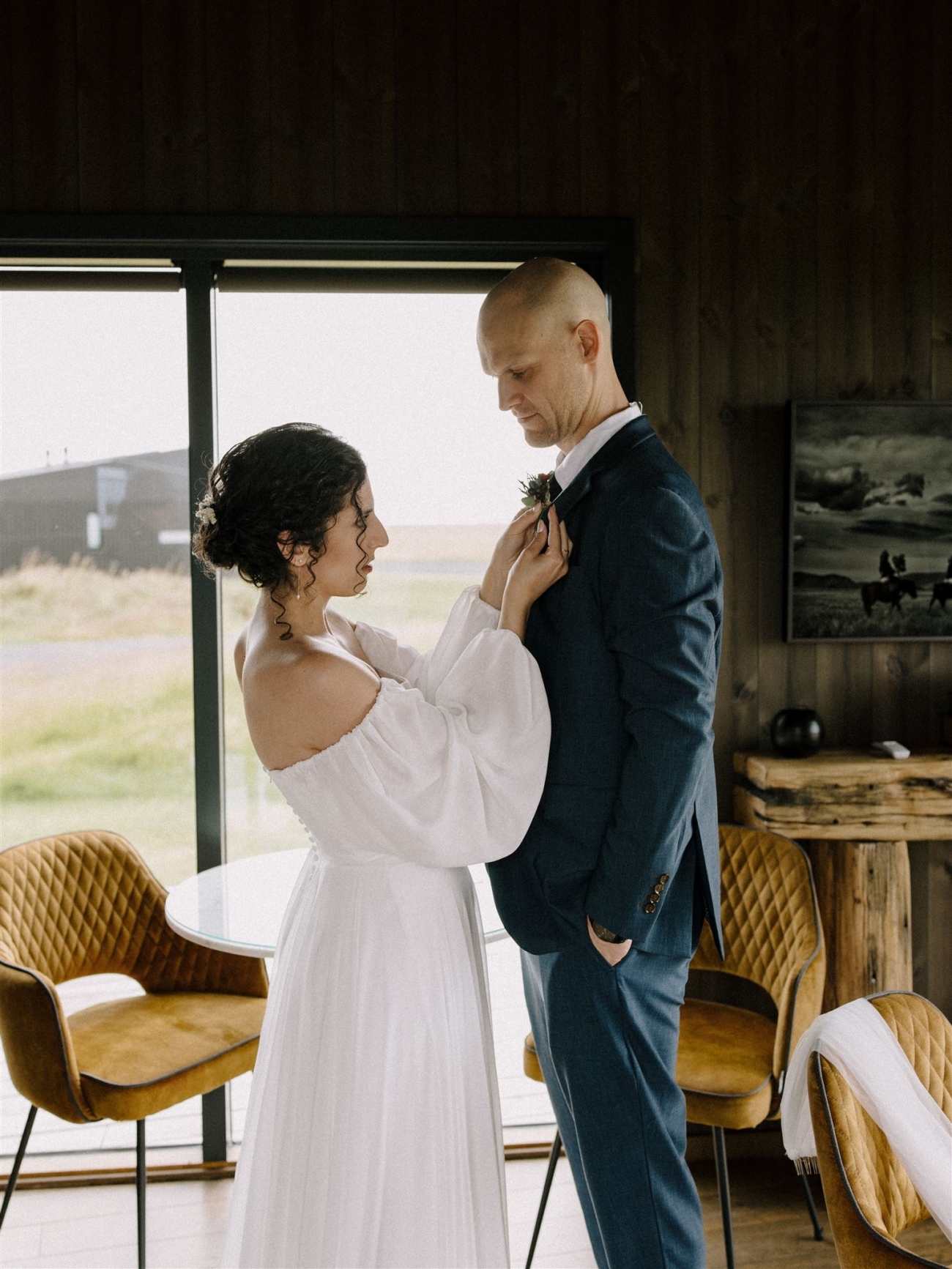 A bride fastens the boutonniere on a groom as they get ready together in a cabin at the start of their Iceland elopement day.
