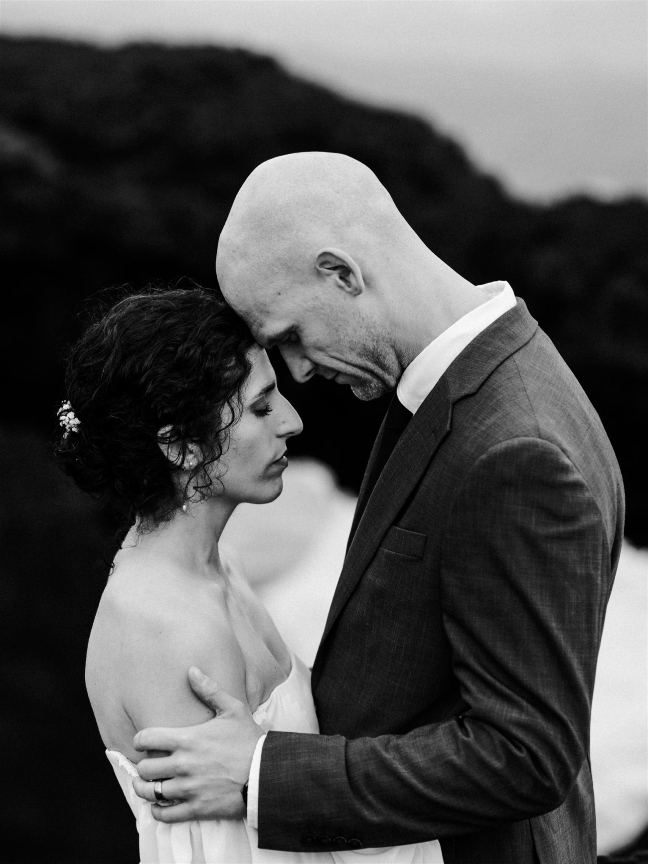 A bride and groom embracing with foreheads touching with beautiful rock formations and the roaring sea behind them on their Iceland elopement day.