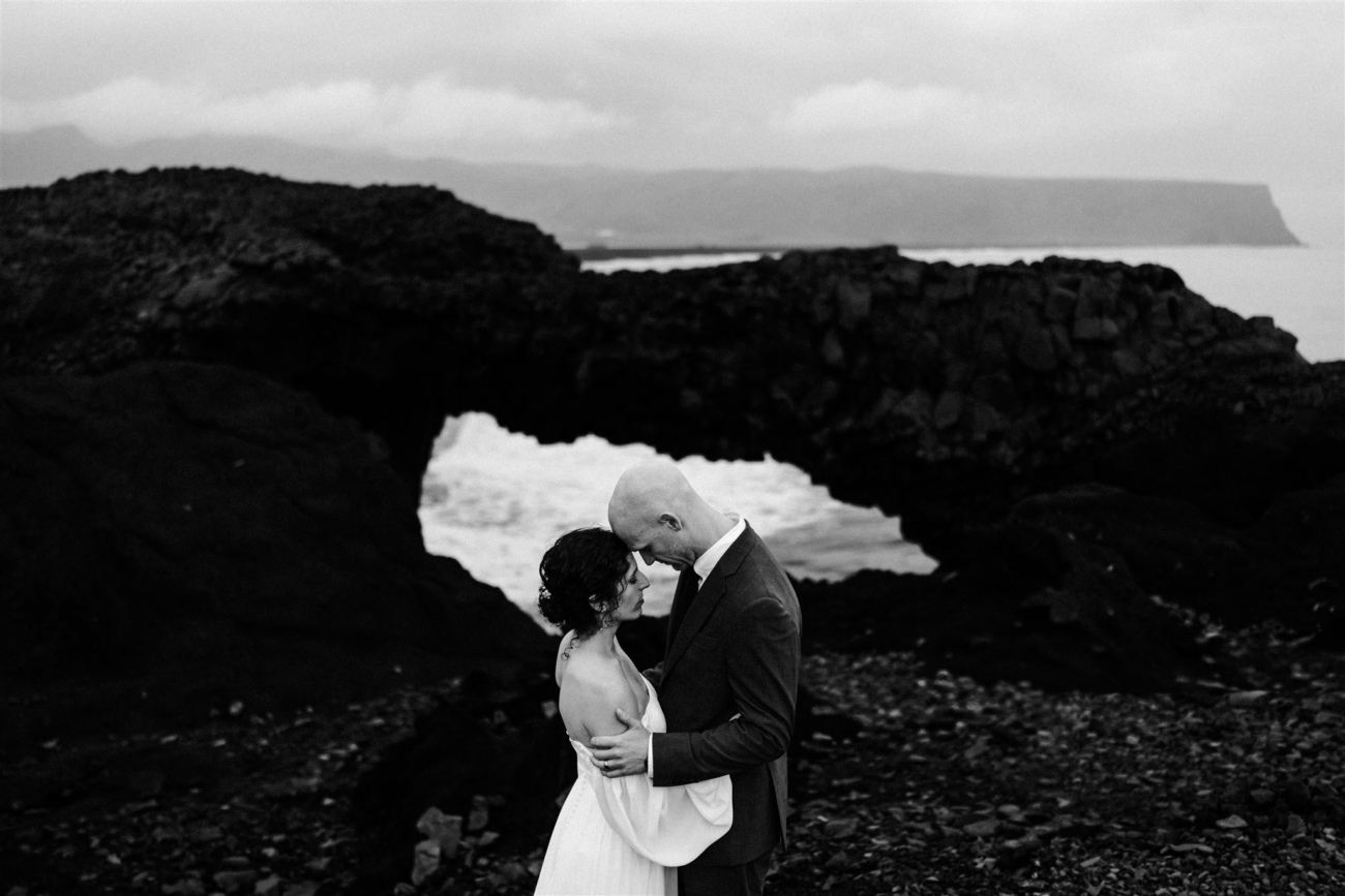 A bride and groom embracing with foreheads touching with beautiful rock formations and the roaring sea behind them on their Iceland elopement day.