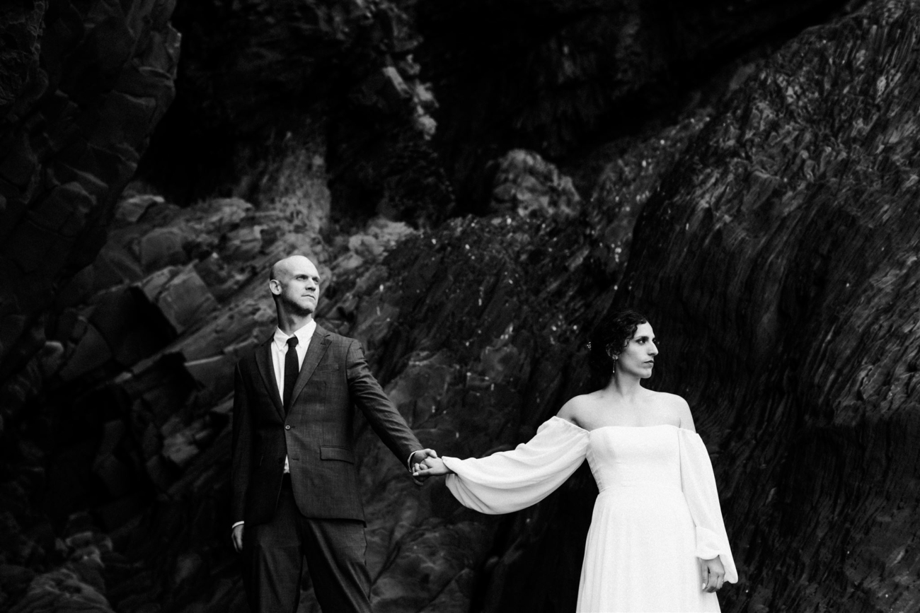A wedding couple holding hands as they walk along the basalt bones of the Reynisfjara black sand beach on their Iceland elopement day.