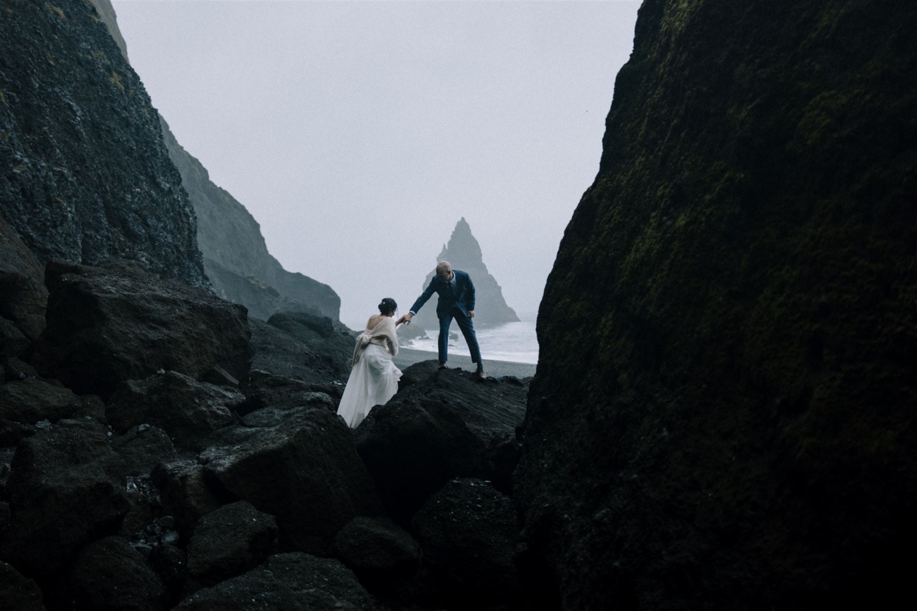 A groom helps his bride climb a big rock on Reynisfjara black sand beach while eloping in Iceland.