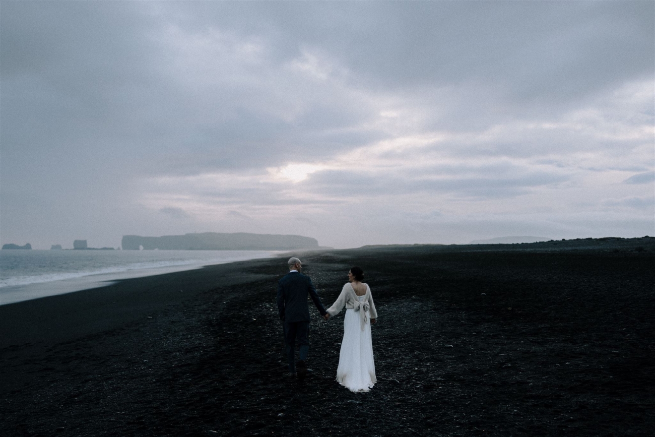 A wedding couple walks into a gloomy sunset on a black sand beach at the end of their Iceland elopement day.