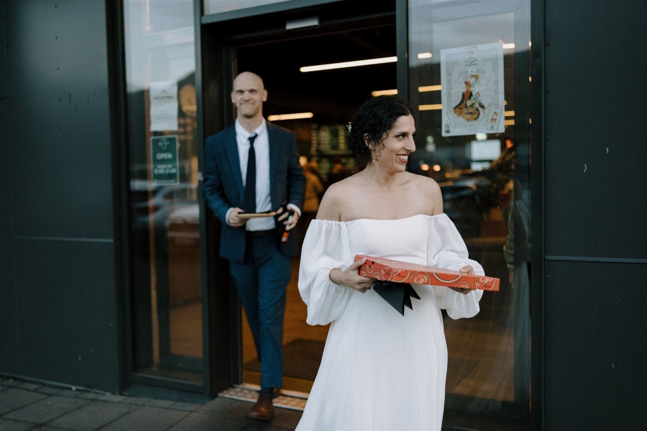 A bride and groom exit a pizza place with a pizza box in Vík, Iceland on their Iceland elopement.
