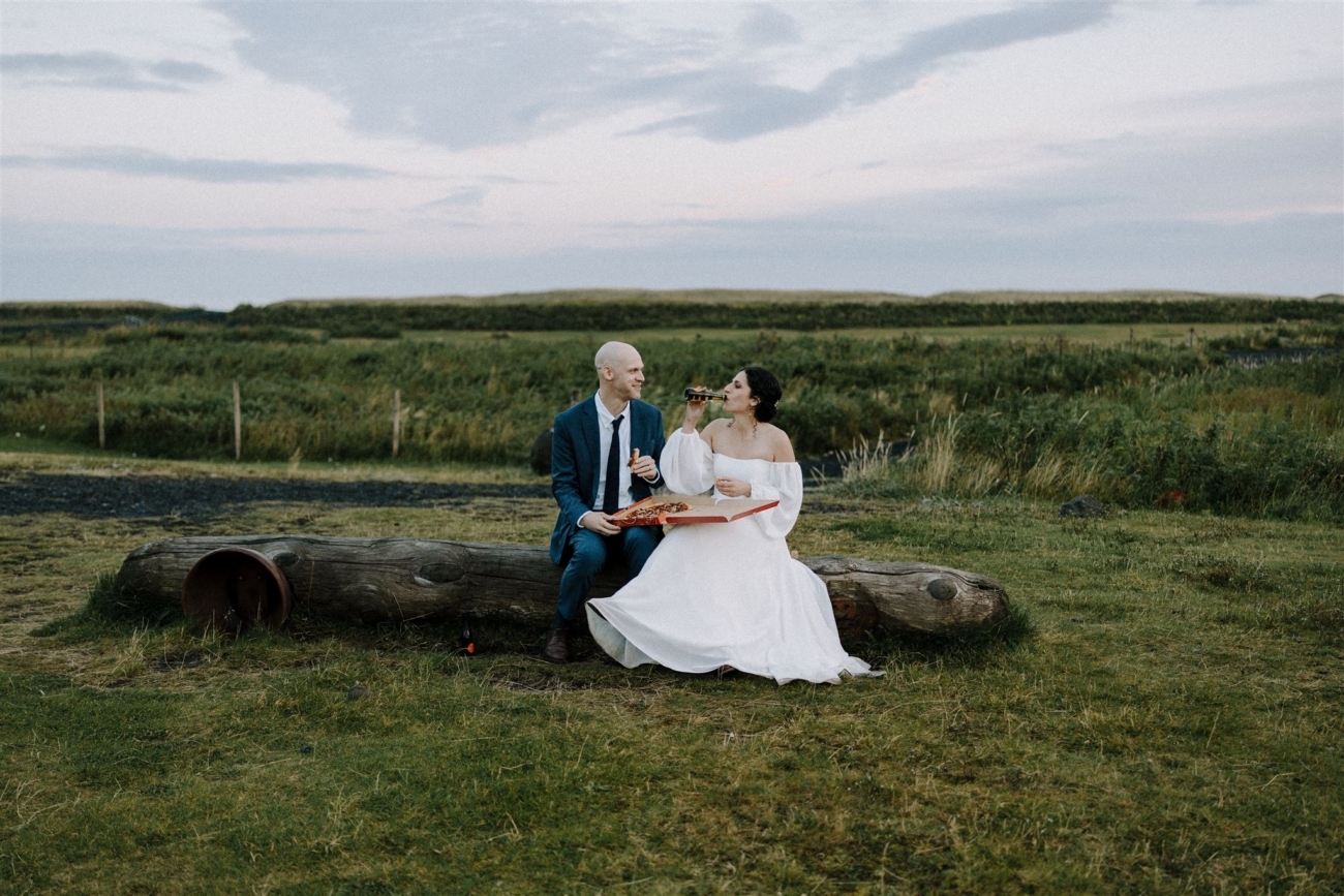 A bride and groom enjoy some prosecco and pizza sitting on a driftwood log at the end of their Iceland elopement.