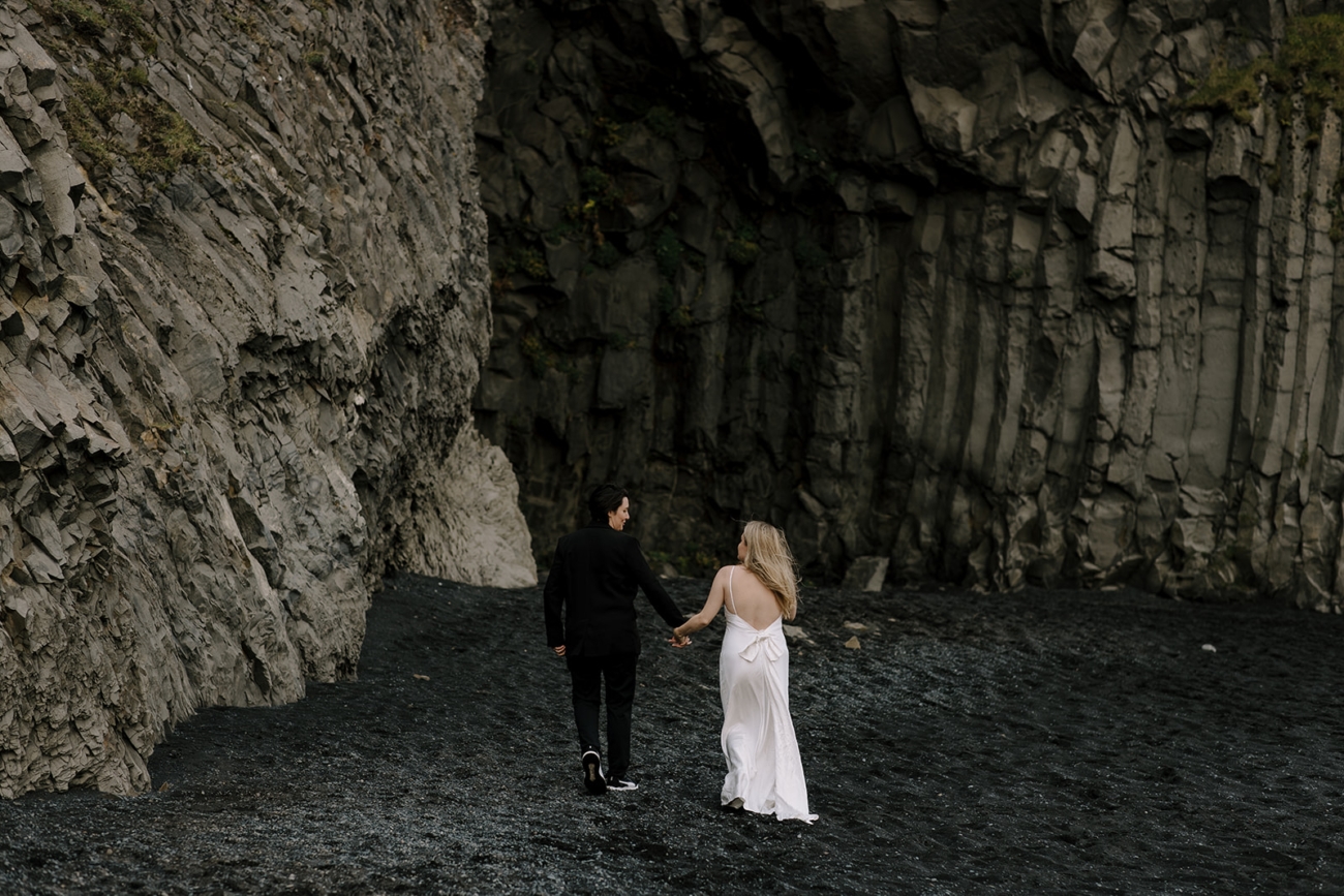 A same sex couple walks away from the camera towards rocky walls on a black sand beach.