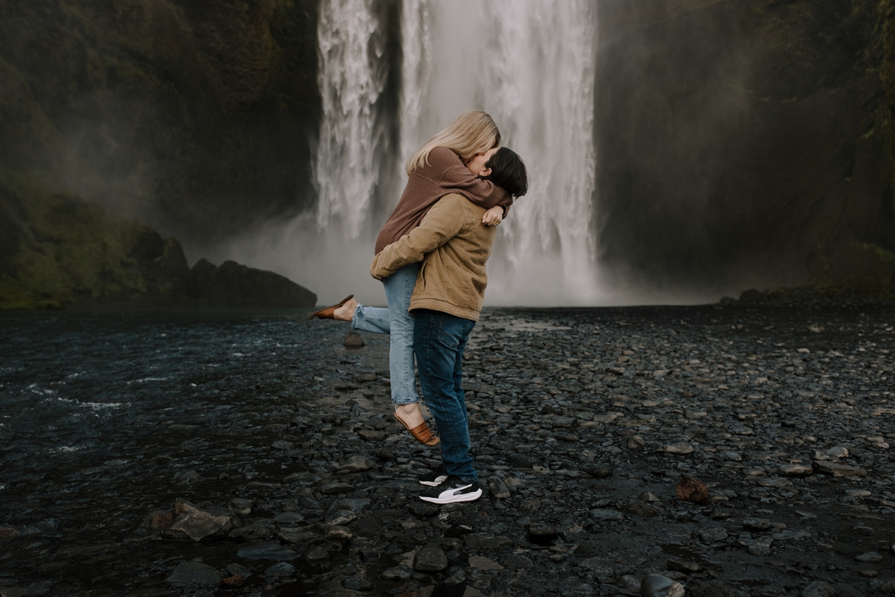 Sara lifting Heather up for a kiss in front of a waterfall.