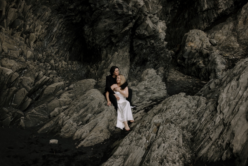 Two women embrace on a jagged rock formation at the black sand beach in Iceland during their Iceland engagement photoshoot.
