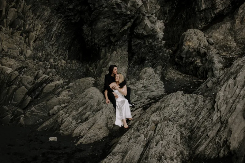 Two women embrace on a jagged rock formation at the black sand beach in Iceland during their Iceland engagement photoshoot.