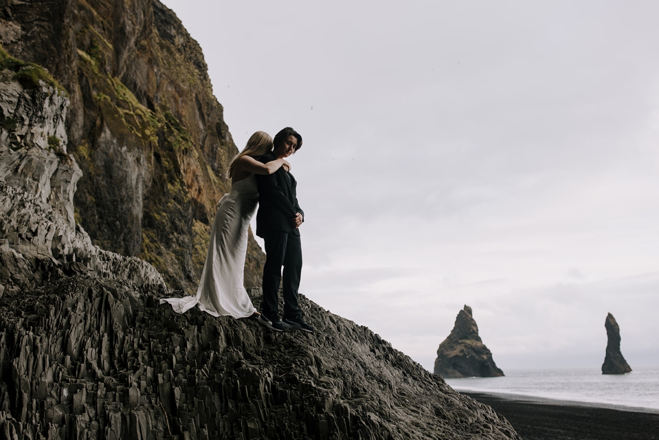 The couple standing on a high rock overlooking the beach and sea stack in the background.