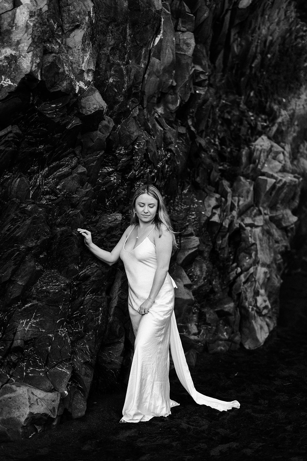 A solo portrait of a women in a white flowy dress on a black sand beach, standing in front of a patterned rock wall.