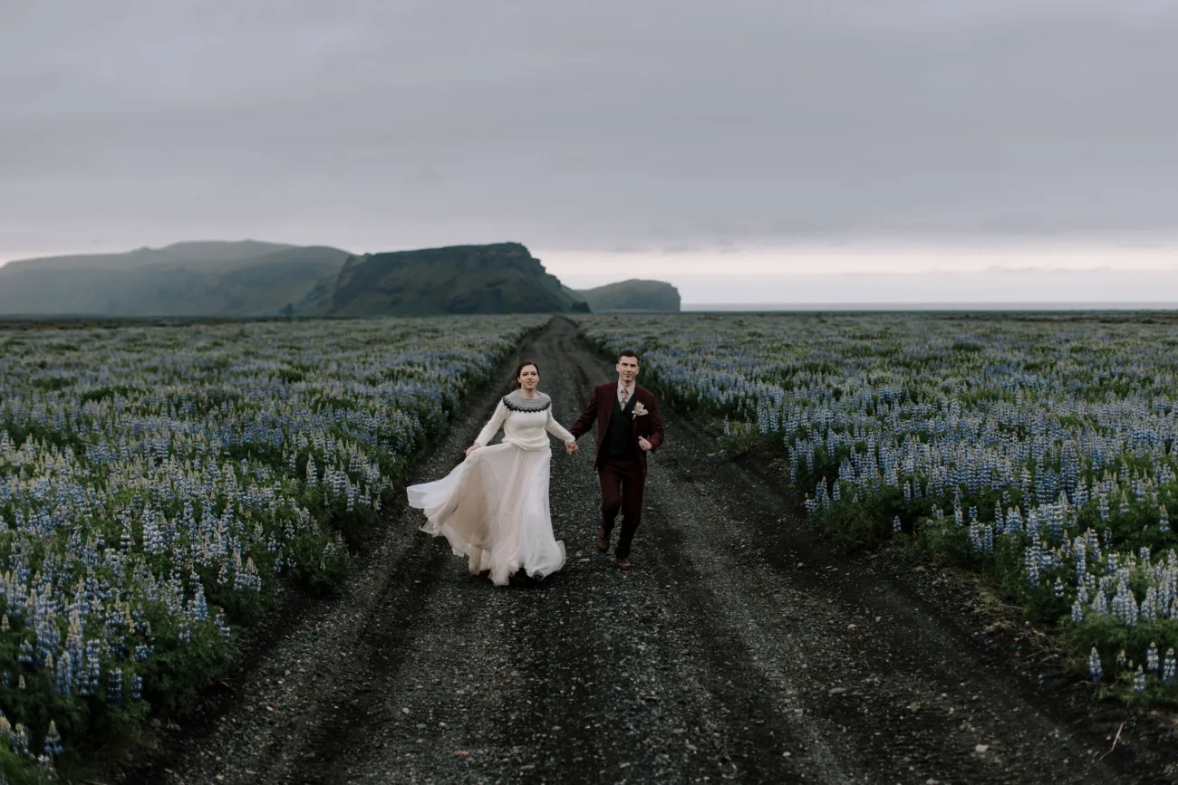 Bride and groom running through lupin field, bride wearing a wool sweater