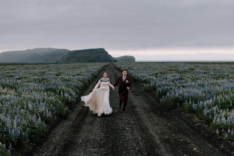 Bride and groom running through lupin field, bride wearing a wool sweater