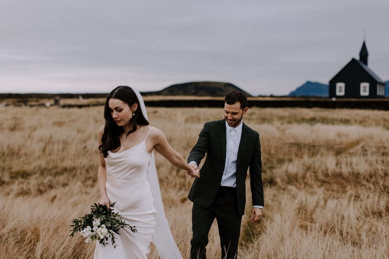 A couple walks in the golden fields behind the black church in Iceland.