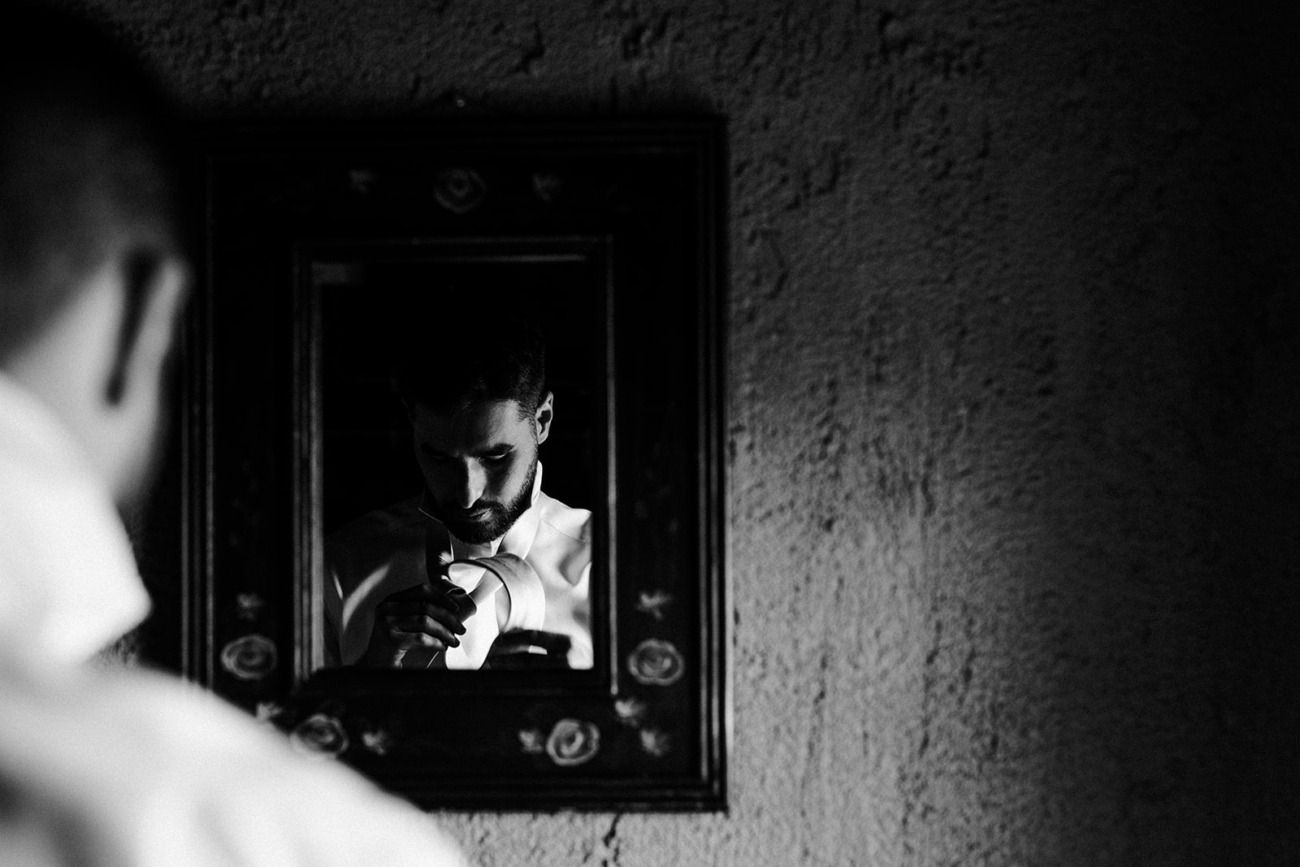 A groom fixes his tie in a wall hanging mirror in a dark little cabin. 