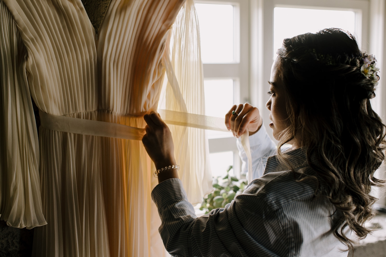 A bride gets her dress ready as it hangs in front of a window with soft light shining through the fabric. 