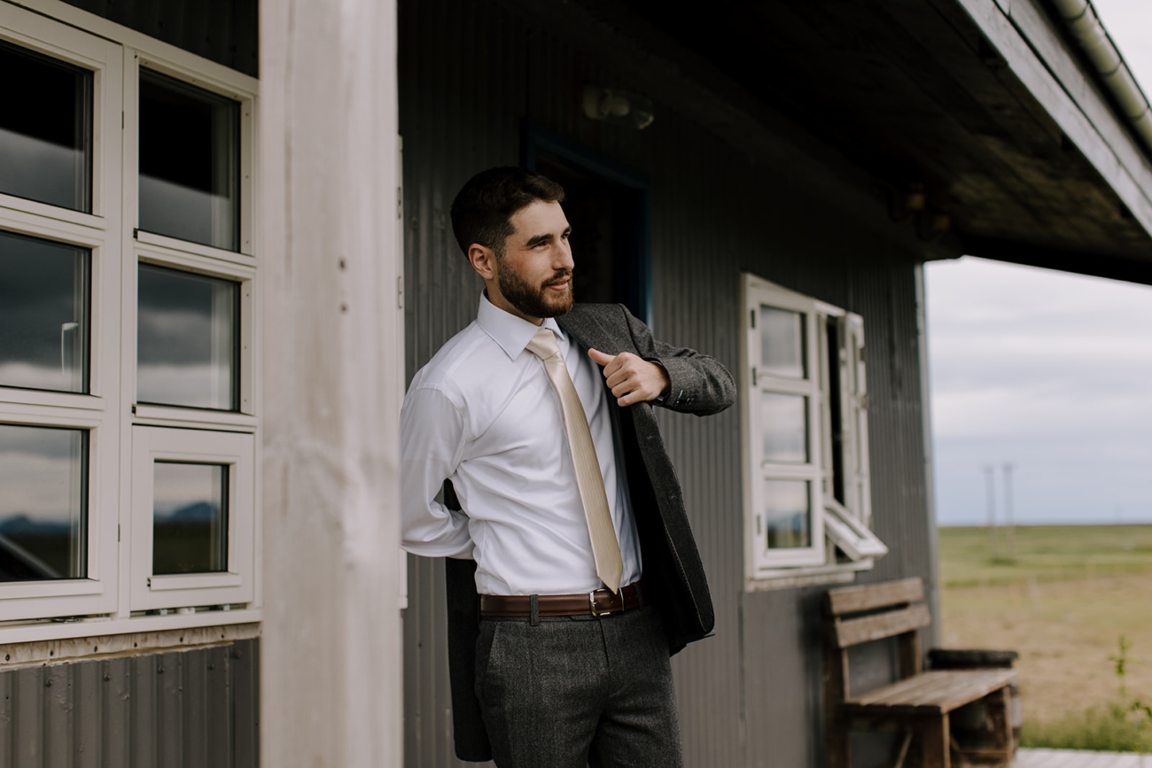 A groom puts on his jacket in front of a little rural cabin. 