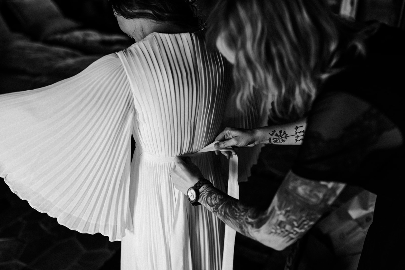 A bride gets her hair and make up artist to button up her dress before she walks out to the porch to meet her husband to be. 