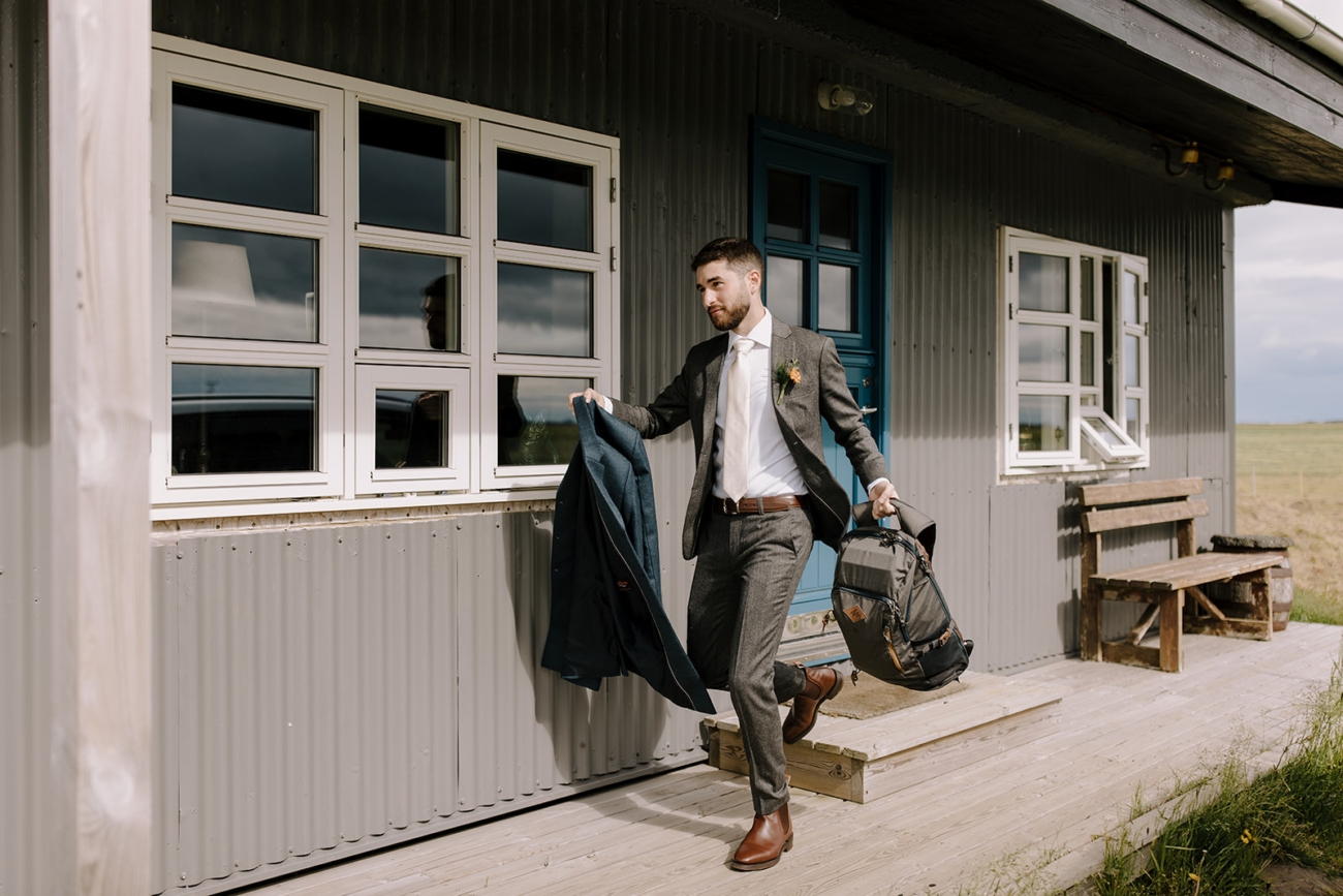 A groom runs with a backpack and an overcoat in is hands towards is elopement day in Iceland. 