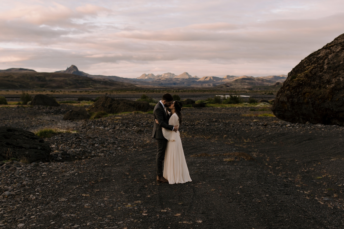 A couple in wedding attire embrace and kiss in a mountain hall in the last light of the midnight sun in Iceland.