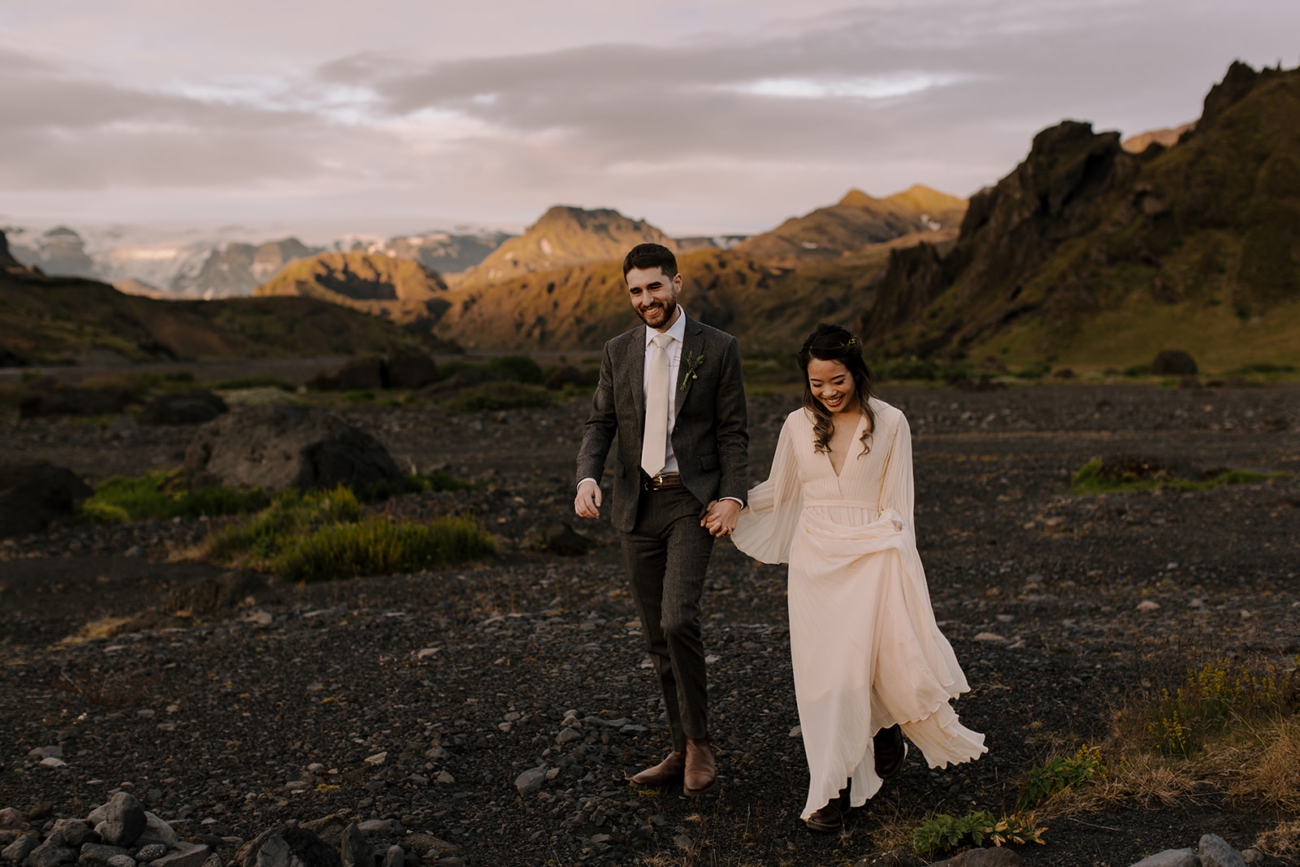 A bride and groom walk hand in hand with huge grins on their faces in a vast landscape in the highlands of Iceland.