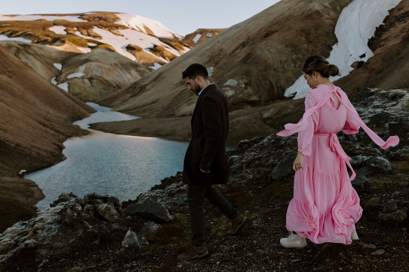 A couple hikes towards an alpine lake as the wind rustles their clothes in the mountains of Iceland.