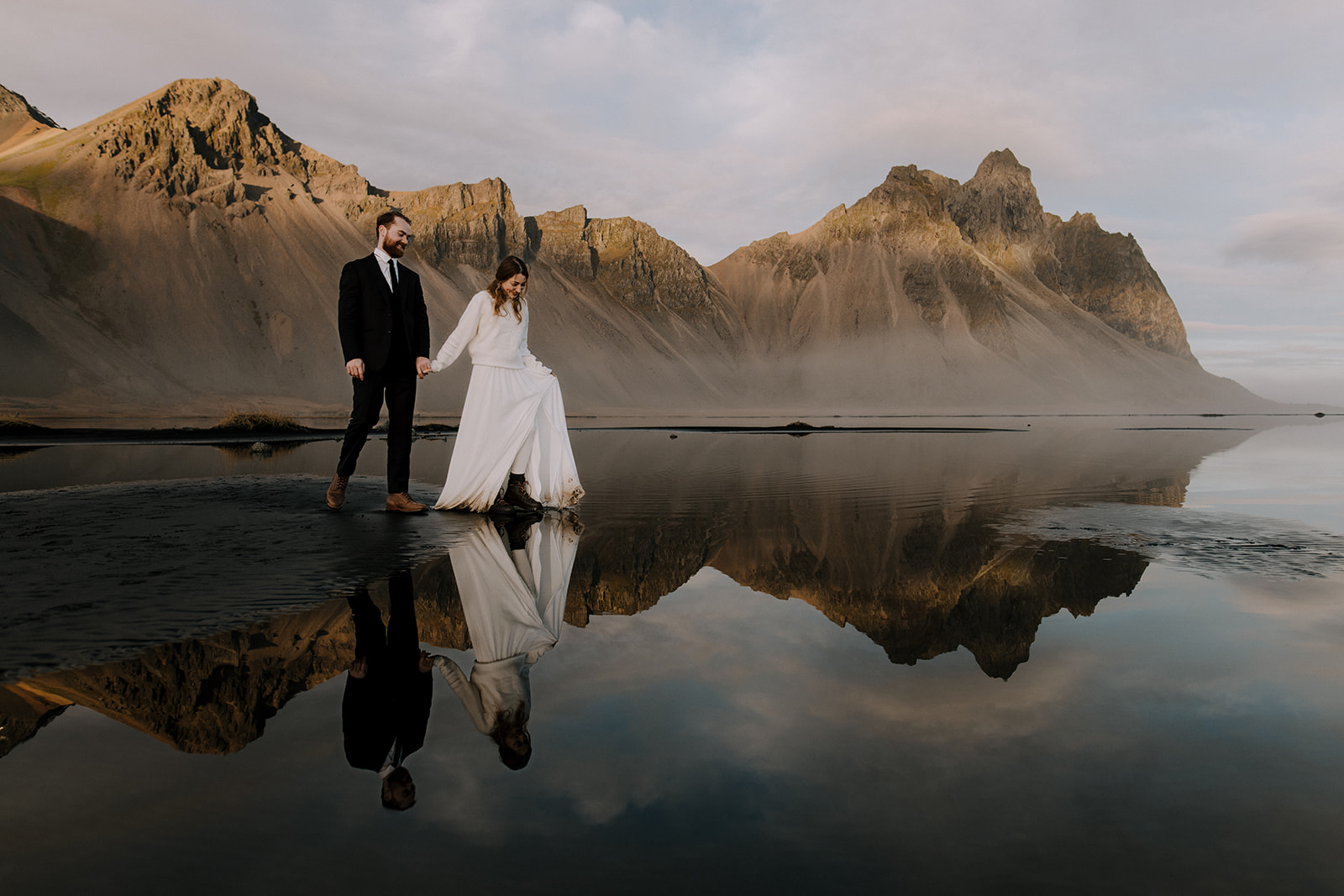 A couple taking a stroll on wet black sand that mirrors it's surroundings perfectly. Creating a surreal atmosphere.