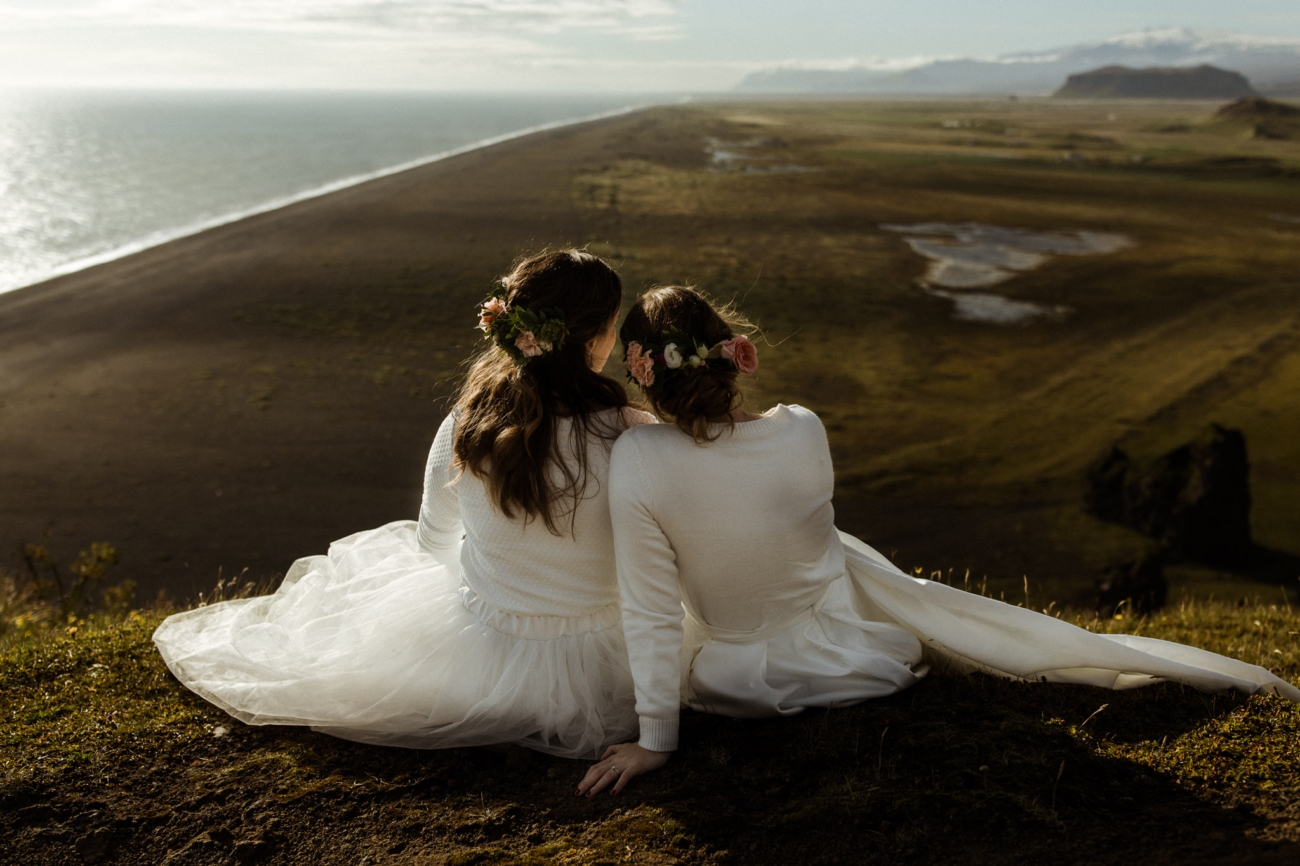 A lesbian couple sits in their wedding dress on top of a cliff on a summer day in Iceland admiring the black sand beach view below them.