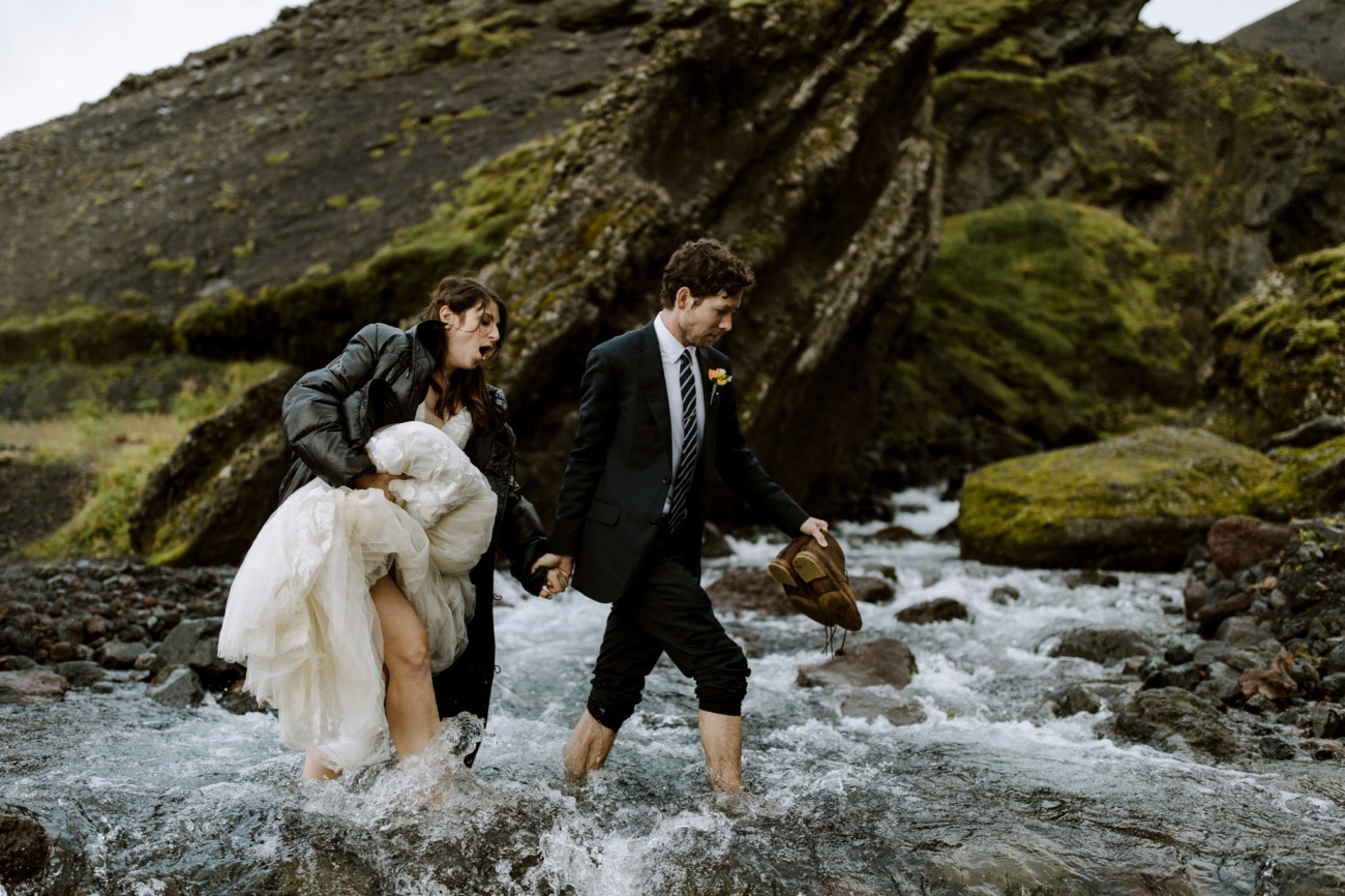 A couple wades over a freezing river in their wedding outfits on their Iceland elopement in the highlands.